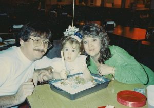 1 year old with her hands in her birthday cake. Parents on both sides. 